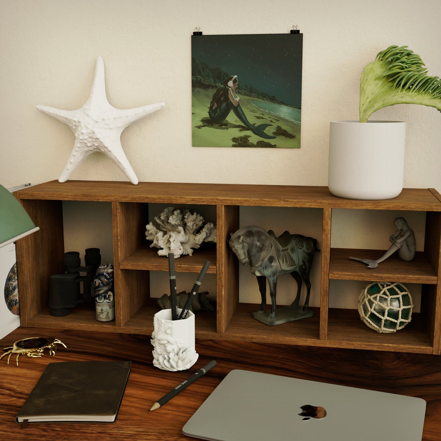 art print of a sad mermaid above wooden shelf and desk with decorative ocean-themed items, books, and a laptop.