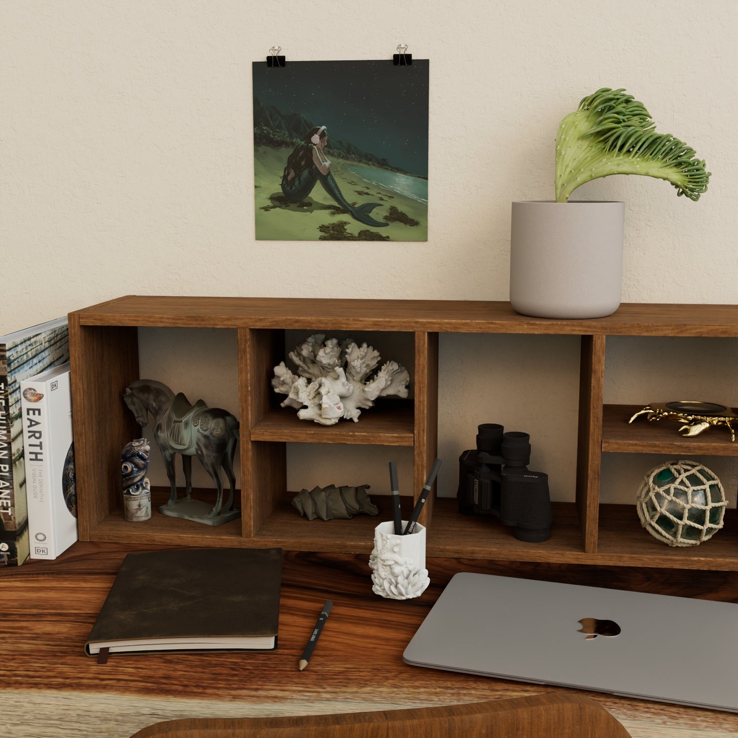 art print of a sad mermaid above wooden shelf and desk with decorative ocean-themed items, books, and a laptop.