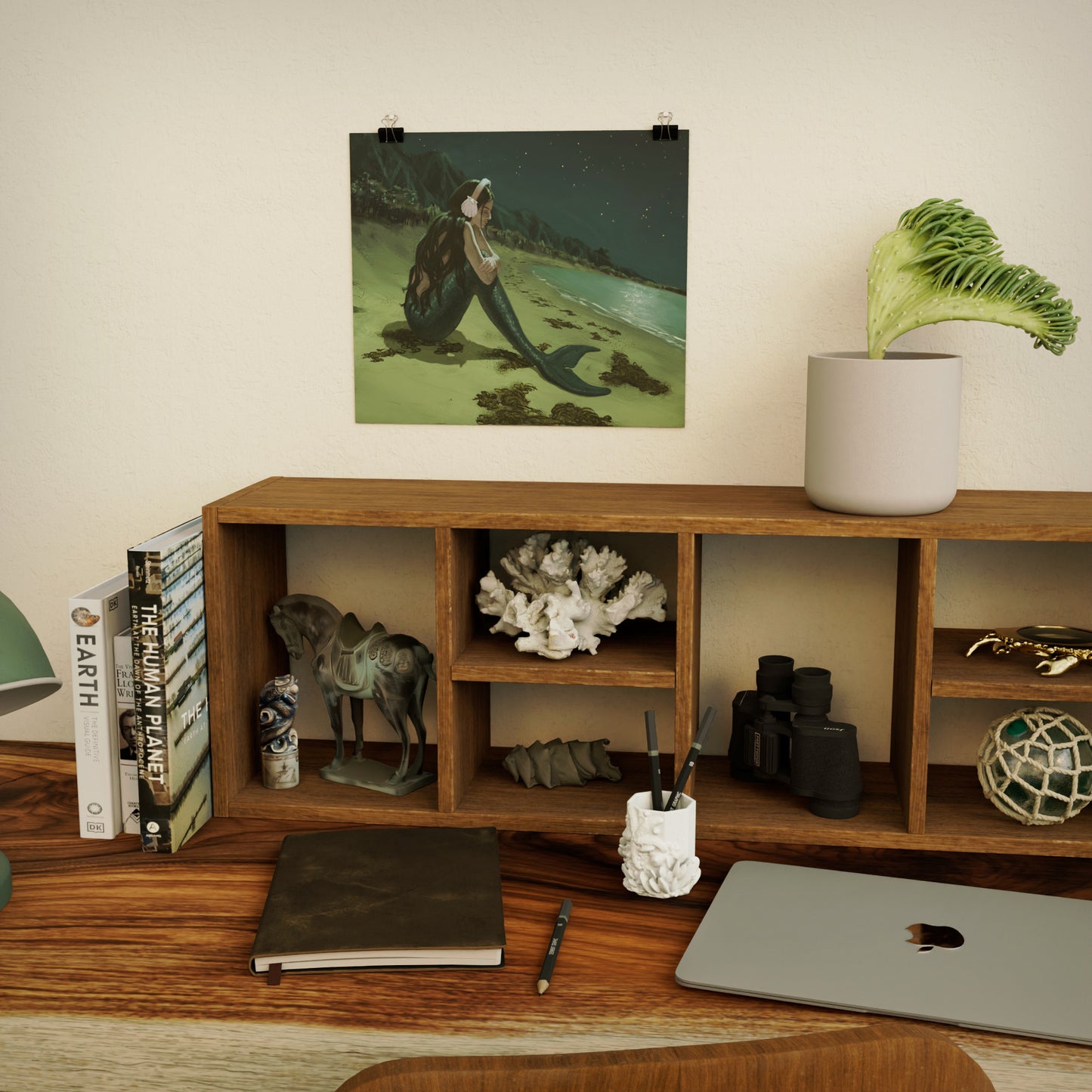 art print of a sad mermaid above wooden shelf and desk with decorative ocean-themed items, books, and a laptop.